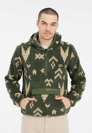Man draagt een groene en beige fleece trui met patroon, half-ritssluiting en voorzak met label "Protest High Performance".