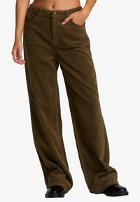 Person wearing loose, brown corduroy pants with hands in pockets and black shoes, shown from mid-torso to feet on a white background.