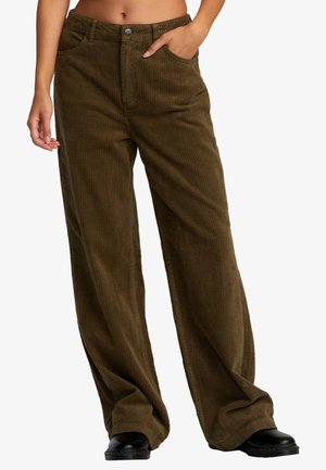 Persona che indossa pantaloni di velluto a coste marroni larghi, con le mani nelle tasche e scarpe nere, mostrata dalla parte centrale del torace fino ai piedi su uno sfondo bianco.