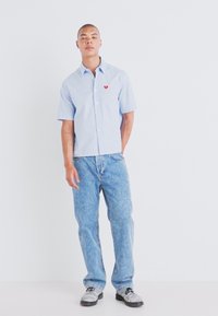 Chemise bleu clair à manches courtes avec un logo cœur rouge, associée à un jean bleu délavé et des chaussures grises. Design simple et décontracté.