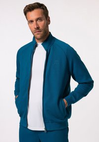 Sudadera con cremallera en color teal, cuenta con un alto cuello y puños acanalados, con un sutil logo en el lado izquierdo. Tela suave y dos bolsillos frontales.