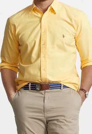 Mann trägt ein gelbes Hemd mit Knöpfen und hochgekrempelten Ärmeln, beige Hose, blauen gestreiften Gürtel und eine Armbanduhr, Hände in den Taschen.