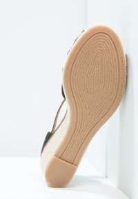 Semelle de sandale avec dessous en caoutchouc couleur beige et grip texturé, talon compensé avec texture tissée, et brides noires visibles sur le côté.