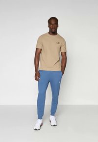 Beige t-shirt med kort ärm och logotyp på bröstet, tillsammans med ljusblå avsmalnande joggers och vita sneakers med svarta detaljer.