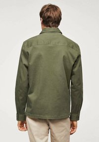 Vihreä puuvillapaita, jossa on kaulus, pitkät hihat ja suora helma. Takana on olkasauma ja nepparit hihansuissa.