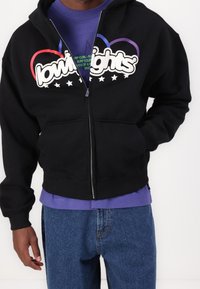 Sweat à capuche noir zippé avec un logo graphique, comprenant des accents rouges, violets et blancs, ainsi qu'une poche kangourou. Porté par-dessus une chemise violette.