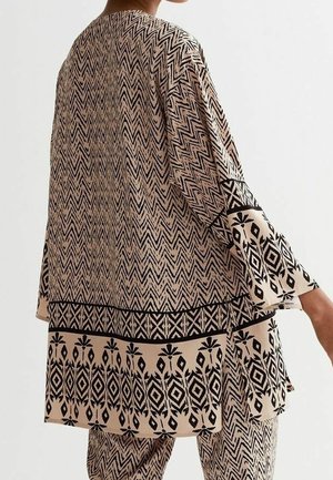 Løsbluse i beige stof med sorte geometriske mønstre. Inkluderer brede ærmer og en kant med indviklede detaljer.