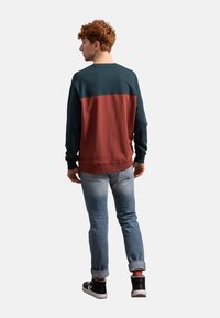 Kleurblok sweatshirt in donkergroen en roestkleur met een geribbelde rand, geplaatst tegen een grijze achtergrond. Gepaard met lichtblauwe jeans en zwarte sneakers.