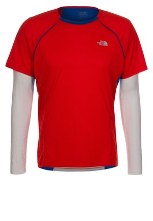 Chemise rouge à manches courtes portée par-dessus un vêtement blanc à manches longues, avec le logo The North Face sur la poitrine gauche et des détails bleus à l'intérieur du col.