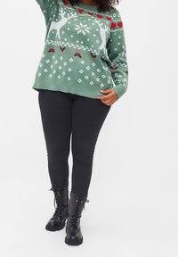 Grüner Strickpullover mit weißen Rentieren und roten Herzen. Verfügt über ein gemustertes Design mit Rauten und einem gerippten Saum. Kombiniert mit schwarzen Jeans und Stiefeln.