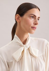 Blouse crème avec un col à nouer, fabriquée en tissu léger. Présente des détails plissés et une texture lisse, adaptée à diverses occasions.