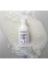 Redken Acidic Bonding Concentrate Shampoo in einer weißen Flasche mit Klappdeckel. Das Etikett zeigt farbenfrohe Akzente und Informationen zum 7% Bonding-Komplex. Blasen umgeben die Flasche.