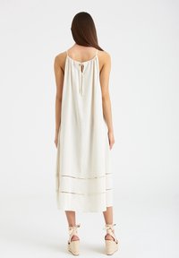 Ärmelloses beiges Kleid mit einer gerafften Rückenschnürung, das horizontale Nähte aufweist. Getragen mit hellen Keilabsätzen.