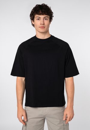 Young man with curly hair wearing a plain black oversized short-sleeve shirt and light beige cargo pants, standing against a white background.