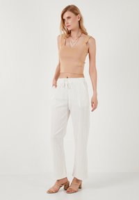 Top cropped de punto beige, pantalones blancos con cordón y un acabado texturizado, y sandalias de tacón cuadrado en tono marrón. La modelo posa contra un fondo liso.