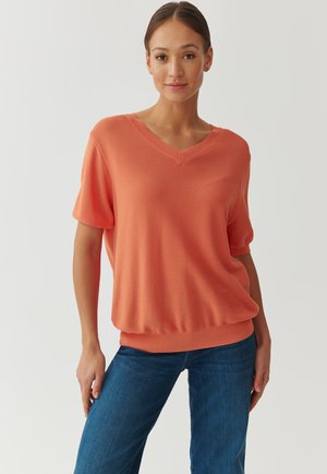 Korallenfarbener V-Ausschnitt-Pullover mit kurzen Ärmeln, gerippter Textur, lockerer Passform und geripptem Saum, kombiniert mit blauen Jeans.