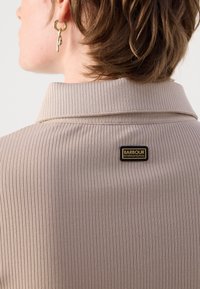 Beiger gerippter Stoffkragen mit detaillierter Stickerei, auf der Rückseite mit einem schwarzen Etikett, das "BARBOUR INTERNATIONAL" in Gold zeigt.