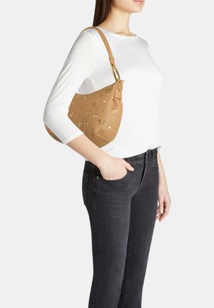 Donna che indossa un top bianco e jeans neri porta una borsa a tracolla in pelle color cuoio con decorazioni dorate a forma di farfalla e cuore.