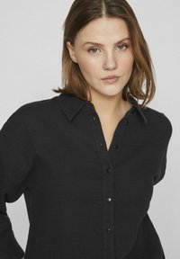 Črna srajca s ovratnikom iz teksturirane tkanine, z gumbom spredaj in zavihanimi rokavi. Vidna je subtilna karirana vzorec.