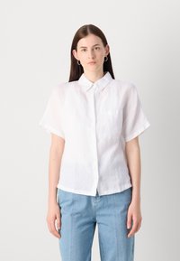 Camisa de linho branca de manga curta com colarinho, fecho frontal por botões, e um pequeno bolso no lado esquerdo do peito, combinada com calças de ganga azul claro.