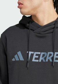 Siva trenirka s prednjim prominetnim modrim grafičnim logom, s kapuco na zatezanje in mehko teksturo. Dopolnjena z črnimi zatezniki.