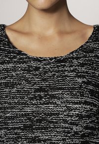 Frau trägt ein schwarzes und weißes strukturiertes Strickoberteil mit einem weiten, runden Ausschnitt, der den Hals und oberen Brustbereich zeigt.