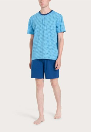 Uomo in piedi a piedi nudi che indossa una camicia azzurra a maniche corte con motivo e bottoni e pantaloncini blu scuro su uno sfondo semplice.