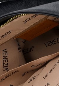 Interiør af en sort læderhandtaske med lys brunt stofforing. "VENEZIA" trykt mønster overalt og et mærke med præget tekst. Guld lynlås kant.