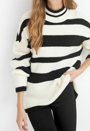 Pullover a righe bianche e nere con collo alto, lavorazione a maglia testurizzata e polsini a coste. Indossato con pantaloni neri, che mettono in evidenza una vestibilità rilassata.