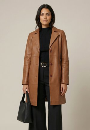 Kvinde iført en brun læderfrakke over sort outfit, holder en sort håndtaske med neutralt udtryk på en ensfarvet baggrund.