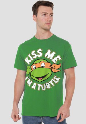 Grünes Baumwoll-T-Shirt mit einem Cartoon-Schildkröten-Gesicht und einem orangefarbenen Bandana, auf dem steht "KISS ME I'M A TURTLE" mit auffälligen weißen und gelben Akzenten.