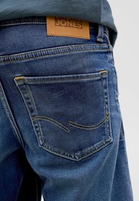 Jean en denim bleu avec une coupe classique. Présente une étiquette en cuir marron et des coutures jaunes contrastées sur les poches arrière.
