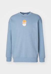 Sudadera azul claro con cuello redondo y mangas largas. Presenta un parche bordado con el logo de Puma en naranja y blanco en la parte delantera.