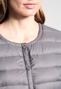 Polstret grå jakke med rund halsudskæring, lodrette sømme og to sorte trykknapper foran. Glat, skinnende tekstur.