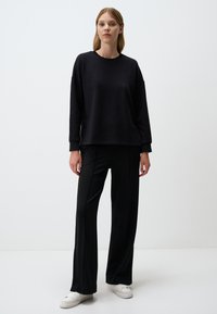 Czarna bluza z długimi rękawami, luźny krój. Połączona z czarnymi spodniami w szerokim kroju i białymi sneakersami, na tle gładkiego białego tła.