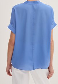 Blusa azzurro chiaro a maniche corte con colletto rotondo, texture morbida e orlo arrotondato. Cucitura visibile lungo il centro della schiena.