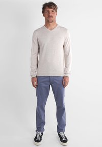Pull beige à manches longues en V, texture légère, associé à un pantalon bleu clair et des baskets noires avec semelles blanches.
