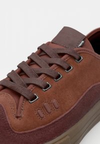 Brązowe zamszowe sneakersy z teksturowanymi sznurówkami, kontrastowymi brązowymi akcentami, metalowymi oczkami i wyściółką kołnierza. Gumowa podeszwa z przezroczystym wykończeniem.