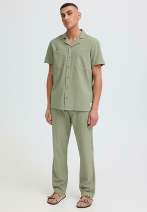 Giovane uomo in piedi che indossa una camicia verde chiaro a maniche corte con bottoni, pantaloni abbinati e sandali beige su uno sfondo semplice.
