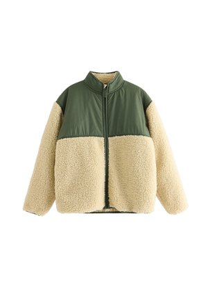 Dreng stående iført en grøn og beige fleece jakke, sorte cargobukser og hvide sneakers, der kigger til siden mod en ensfarvet hvid baggrund.