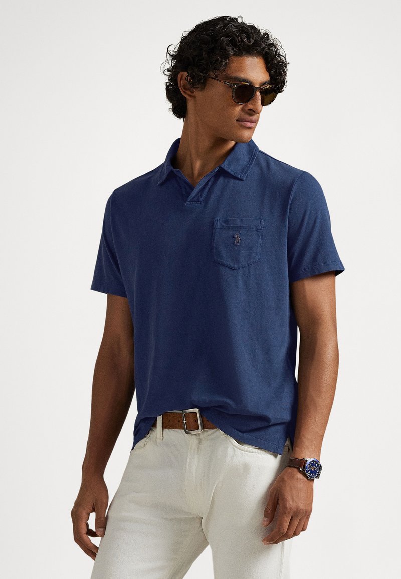 Polo azul-marinho em algodão, com bolso contendo um pequeno emblema de logotipo, combinado com calças de cor clara e um cinto castanho.