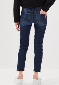 Jean skinny bleu foncé avec une coupe ajustée, comportant des poches arrière avec un subtil détail de couture, associés à des baskets blanches.