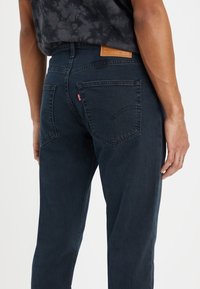 Primer plano de una persona que lleva puestos unos jeans Levi's azul oscuro, mostrando los bolsillos traseros, la etiqueta de cuero de la marca y parte de una camisa negra con estampado.