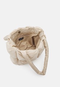 Sac beige rembourré avec une texture douce, de forme ronde, et une bandoulière unique ; il comprend une poche intérieure zippée et une étiquette à l'intérieur.