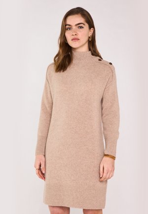 Jeune femme aux cheveux bruns mi-longs portant une robe pull beige avec des détails à boutons sur les épaules et des boucles d'oreilles en créole doré.