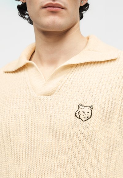 Homme portant un polo en maille beige avec un petit logo de tête de renard brodé sur le côté gauche de la poitrine.