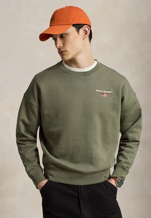 Junger Mann trägt einen olivgrünen Polo Sport Sweatshirt und eine orangefarbene Kappe, Hände in den Taschen, blickt nach links vor einem einfarbigen beigen Hintergrund.