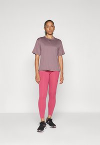 Femeie stand în picioare purtând un tricou sport cu mânecă scurtă în nuanță de mov, colanți roz aprins și adidași negri, pe un fundal alb simplu.