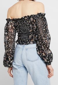 Off-the-shoulder black blouse with floral print, gathered bodice, and long puff sleeves. Back view shows ruched texture and elastic band.