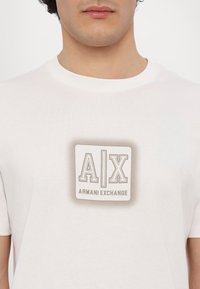 White cotton t-shirt featuring a square graphic with the letters “A|X” and the text “ARMANI EXCHANGE” in a neutral color scheme.